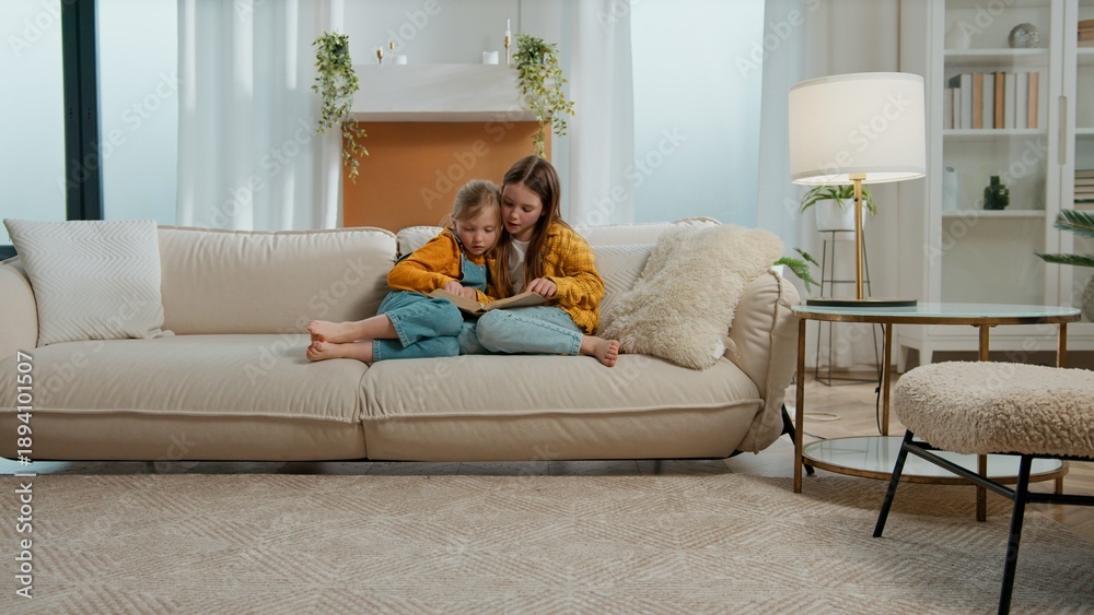 Fototapeta premium Two Caucasian little kids girls sibling children reading book curious sisters read fairy tale watching pictures photos on couch sofa relaxing studying learning together study learn in living room home