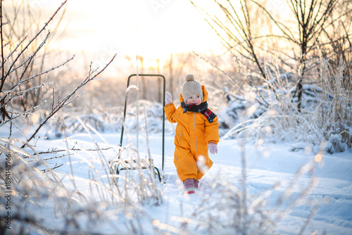 A little girl stands in a bright orange winter suit near a sled - a concept of winter, childhood, winter activities.