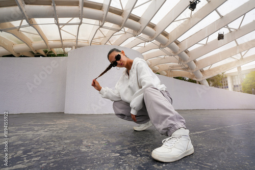 Hispanic woman wearing casual sportswear and sunglasses squatting under a white architectural structure