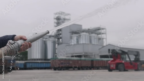 Safety vest, Protective equipment, Tank cars. High-visibility worker sprays pressurized hose while cleaning industrial site beside towering storage silos and freight trains.