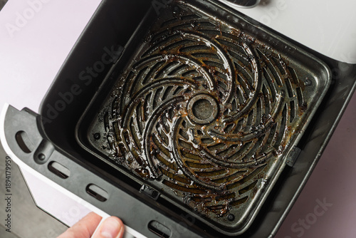 Hand pulls out dirty air fryer basket with burnt grease and food residue, concept of kitchen hygiene, appliance maintenance, cleanup.