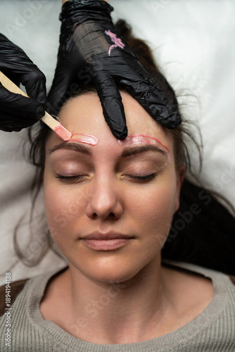 woman receiving eyebrow waxing treatment, gloved esthetician applying warm gel with wooden spatula over brow area, client eyes closed