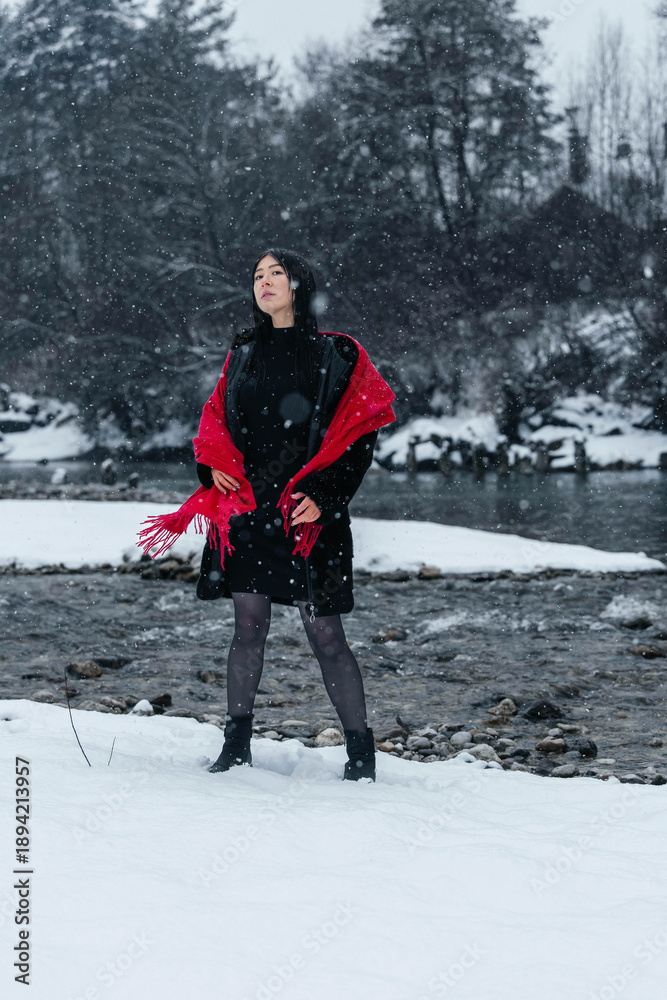 Photography Poster Woman wearing black dress and red shawl stands in snowy landscape near a river, | Wall Art