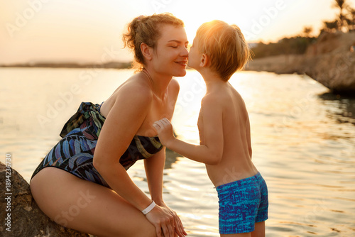 A woman sitting on a rock and a young boy standing in the water, touching noses/kissing. High-contrast sunset background with bright sun flare.