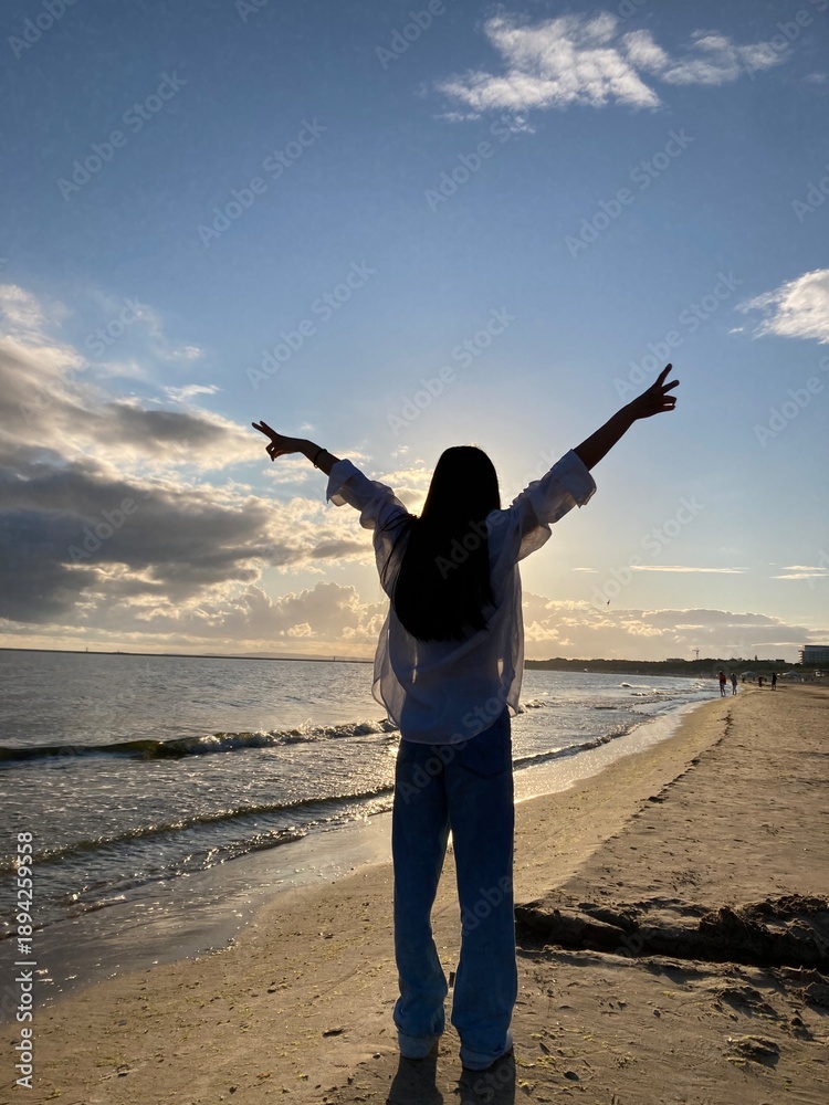 Fototapeta premium Woman Silhouette with Arms Raised on Beach at Sunset