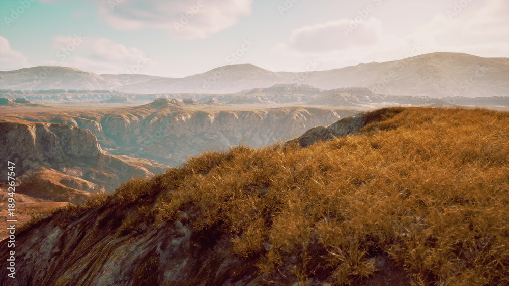 Obraz premium Vibrant grass sways gently atop a rocky ridge as golden light filters through the evening sky, illuminating rugged mountains and valleys in the distance, creating a serene atmosphere of nature.