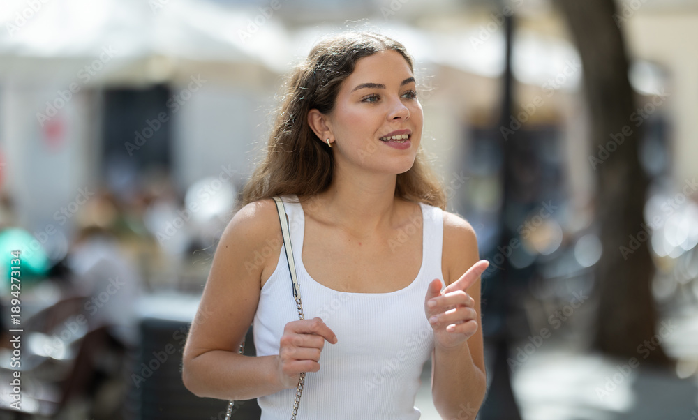 Obraz premium Young woman smiling and walks along sunlit street, walks around city, spends time in public urban space. Person and megalopolis..