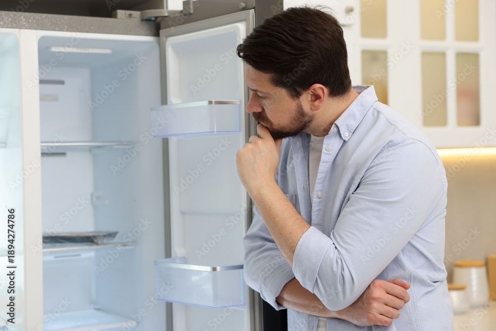 Fototapeta premium Man standing near empty open refrigerator in kitchen
