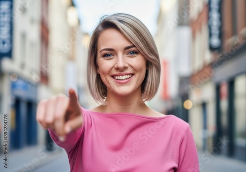 Free Download A smiling young woman with short blonde hair pointing at the camera on a city street with blurred background - GFXLoad