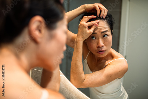 Woman inspecting scalp for hair loss in bathroom mirror