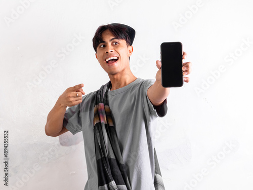 Bild auf Leinwand Excited Indonesian man wearing peci and sarung standing while showing blank smartphone screen mockup isolated on plain background for islamic digital promotion