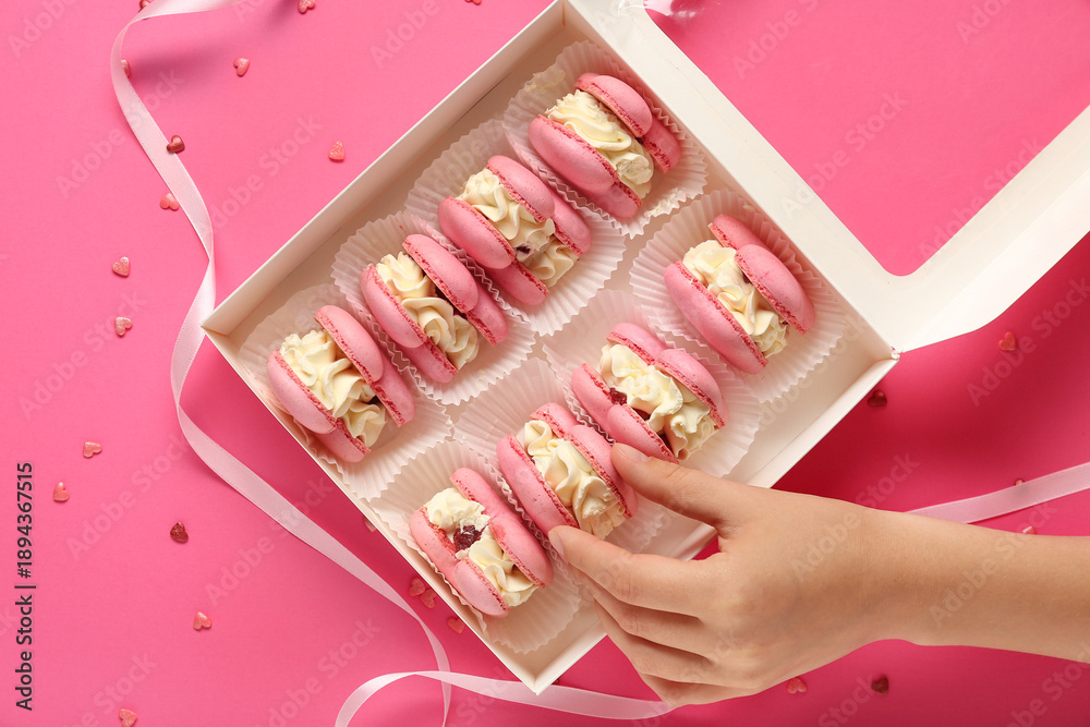 Naklejka premium Female hand with gift box of sweet heart-shaped macaroons on pink background. Valentine's Day celebration