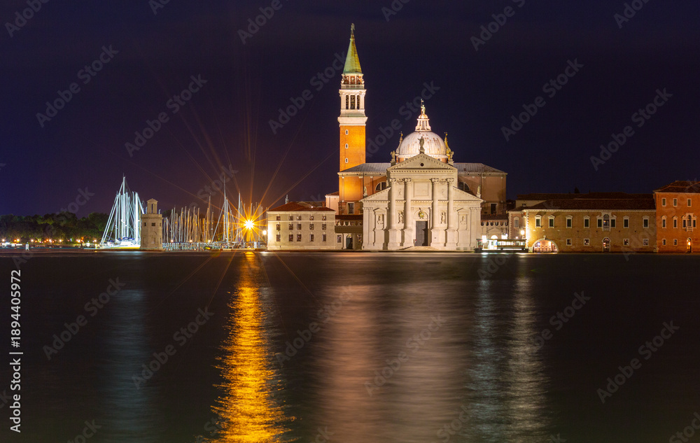 Obraz premium The illuminated facade and bell tower of San Giorgio Maggiore Church in Venice, Italy are reflected in the water under a dark night sky.