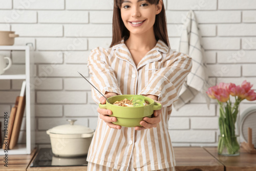 Young woman in pajamas holding bowl with tasty granola and kiwi in kitchen