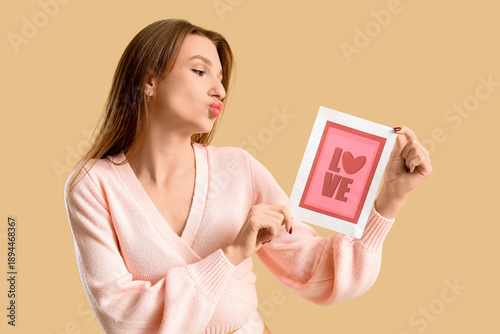 Beautiful young woman holding greeting card with word LOVE and heart on beige background. Valentine's Day celebration