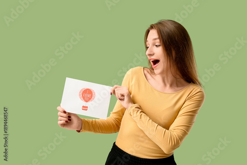 Surprised young woman holding greeting card with text I LOVE YOU on green background. Valentine's Day celebration