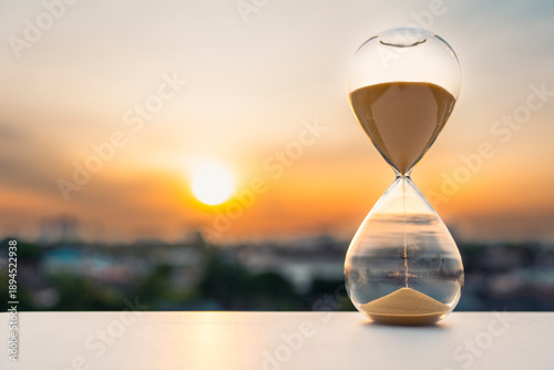 Schilderij op canvas Hourglass on table at sunset with blurred city background symbolizing passing time, limited lifespan, impermanence of life, time management, urgency, reflection, and awareness in a fast-paced modern