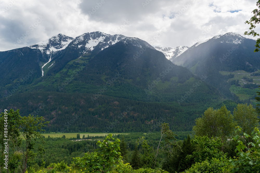 Fototapeta premium Snow-capped mountain peaks with forested slopes above flat river valley.