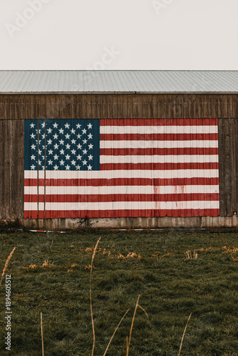 Large American flag painted on the side of a wooden barn. Grass field in the foreground under a grey sky.