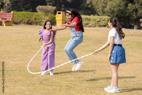 Obraz na plátně Three indian kid friends play skipping rope together in summer park, Children having fun playing outdoor activity