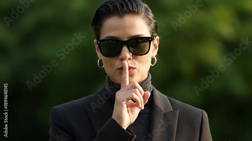medium close up realistic footage anonymous person facing camera silence gesture index finger near lips relaxed confident expression subtle smile elegant posture dark clothing sunglasses natural outdo