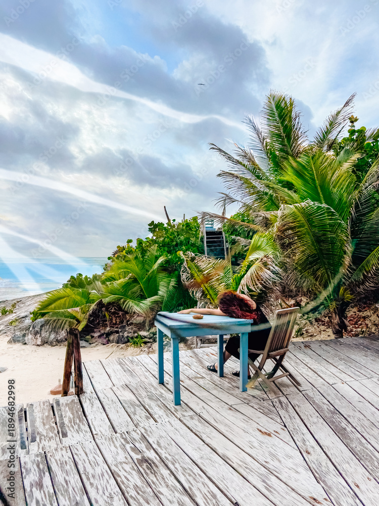 Fototapeta premium Relaxing day at a beachside table with palm trees and sea view. A person sits at a table on a beach deck among palm trees, looking towards the sea in the evening.