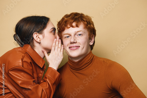 Foto Young woman whispering secret to smiling redhead man with casual clothes, friendship, communication and closeup portrait on beige background with emotions and sharing news