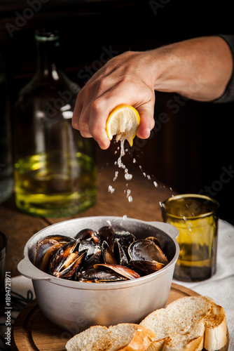 Obraz na plátně Hand squeezing fresh yellow lemon over silver metal pot filled with cooked black mussels seafood dish on wooden table with toasted bread slices and glass bottle of white wine in dark moody background