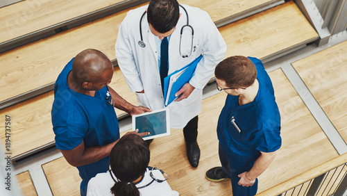 A top-down view of a diverse, multiracial team of four doctors gathered in a huddle. They are wearing blue surgical scrubs under white lab coats. Together, they are reviewing an iPad.