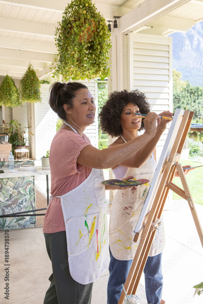 Naklejka premium Asian instructor guiding her student painting on wooden easel at covered porch near plant