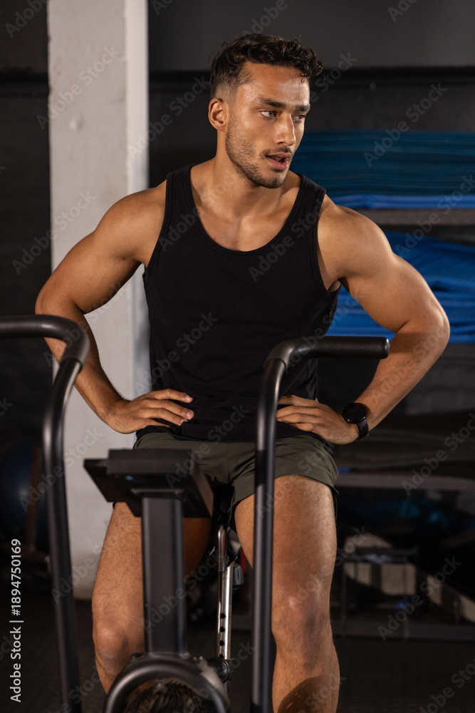 Obraz premium Muscular man pedaling stationary air bike in gym wearing black tank top green shorts and smartwatch