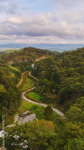 Aerial view of Da Lat suburbs in Vietnam with pine forest, curved roads, and moving vehicles. Landscape showing transportation routes surrounding natural terrain under cloudy sky.