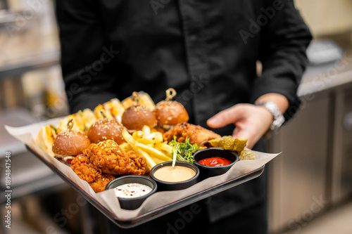 Fotografia A close-up, horizontal, eye-level shot of a server holding a black tray laden with an appetizer platter of mini burgers, fried chicken, french fries, and dipping sauces