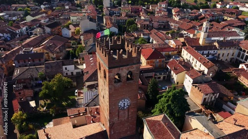 High-angle drone orbiting around a medieval stone clock tower, revealing historic rooftops and dense town architecture in Piedmont.
