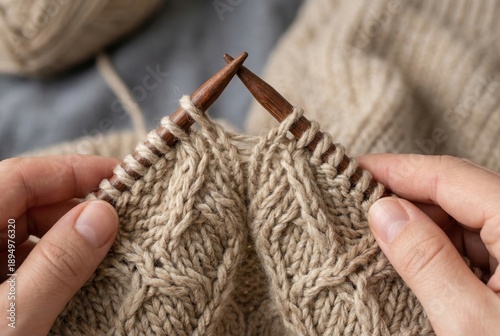 Two hands hold knitting needles as they create a texture with beige yarn. The setting is indoors with natural light. Yarn is nearby, indicating ongoing work