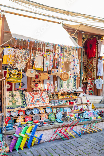 Bright and lively souvenir stall on a charming cobblestone street showcasing handwoven rugs, ceramic plates, beaded ornaments, woven bags, traditional clothing, wooden toys, and decorative crafts