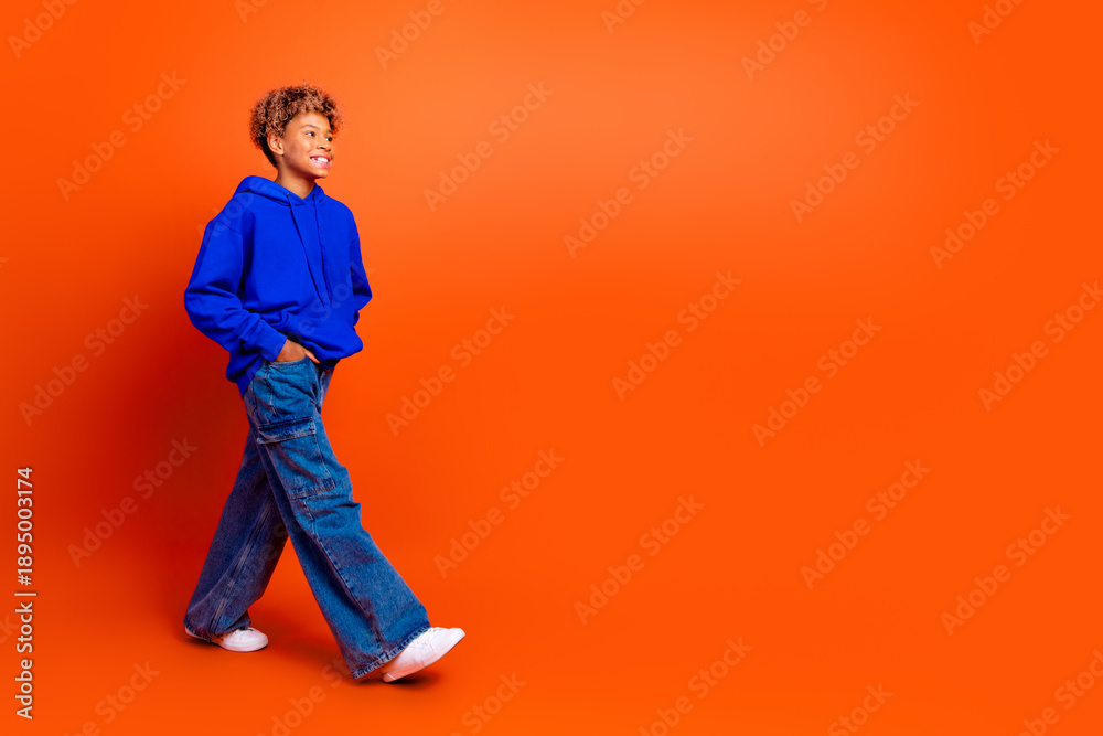 Naklejka premium Young boy in blue hoodie walking on orange background smiling and showing casual fashion lifestyle stock image