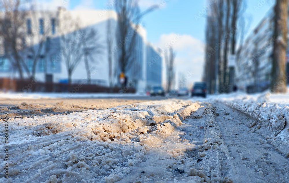 Fototapeta premium Snowy urban street with icy tire tracks and slush, cars driving past apartment buildings on a cold winter day. Low-angle view highlighting winter road conditions