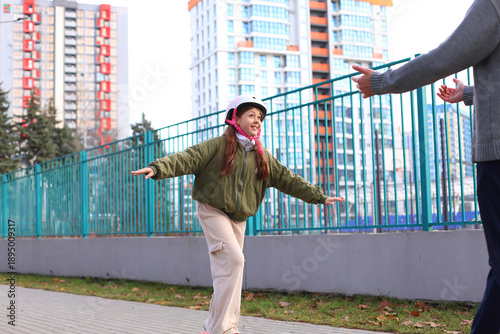 A girl in a helmet balances on a scooter, arms outstretched, as an adult guides her, joyful learning, trust, and supportive coaching in an urban park setting.