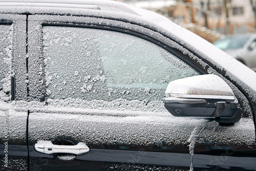 Close up detail of black car window and side mirror completely covered in white frost and hoarfrost patterns on cold winter morning showing extreme freezing weather conditions. Frozen car window