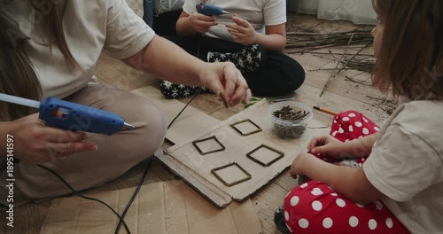 Creative family sitting on the floor making handmade crafts using a hot glue gun and natural materials. Mother helps her children with a diy project, creating decorations from small twigs.