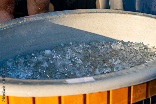 Fényképezés Wooden garden hot tub filled with ice cubes for cold plunge, recovery ice bath for athletes outdoors, ice in blue tub during sunset