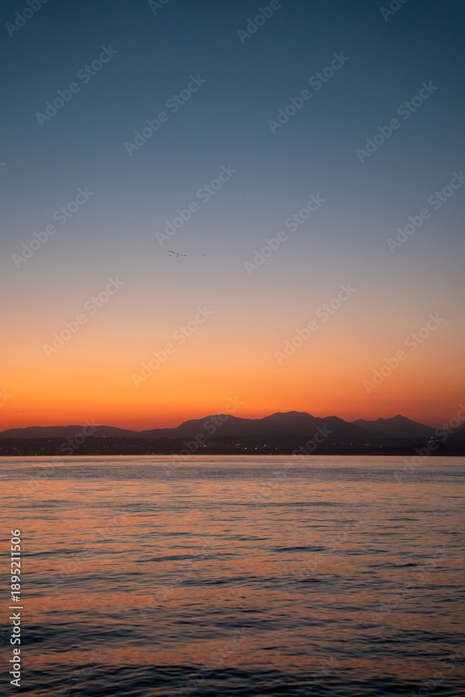 Fototapeta premium Vibrant Orange Sunset over Lake Garda with Mountain Silhouettes and Calm Water