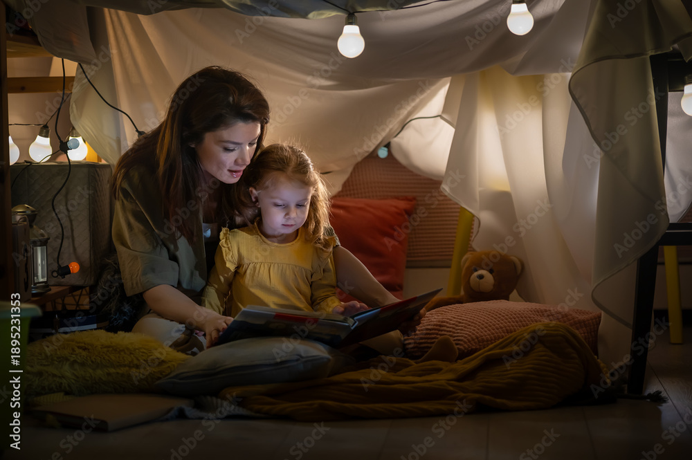 Naklejka premium Mother reading a bedtime story to her young daughter inside a cozy blanket fort at home, glowing lights creating a magical, calm atmosphere of love, comfort, and imagination