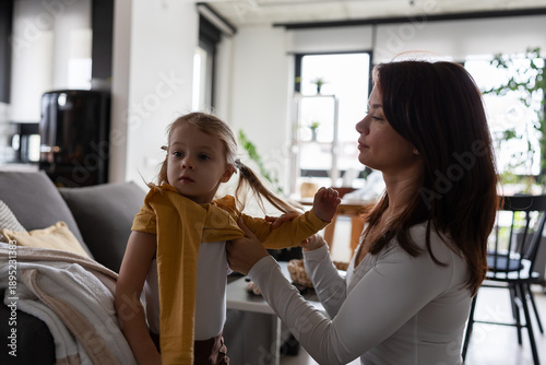 Obraz na plátne Young mother helping her toddler get dressed at home, gently fixing her clothes
