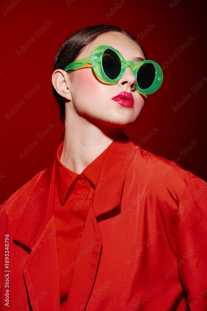 Fototapeta premium Fashion model in bright red shirt and jacket poses with glossy red lips and green sunglasses. Bold makeup complements modern artistic beauty and style studio shot.