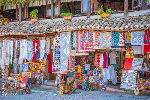 Vibrant street market scene in a historic Albanian old town featuring beautifully handwoven kilim rugs, embroidered fabrics, lace tablecloths, scarves, wooden bowls, ceramic souvenirs, magnets