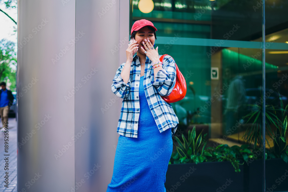 Fototapeta premium Young Asian woman laughing during phone call outdoors, wearing red cap, plaid shirt, blue dress and red bag, standing next to modern glass building, joyful street moment.
