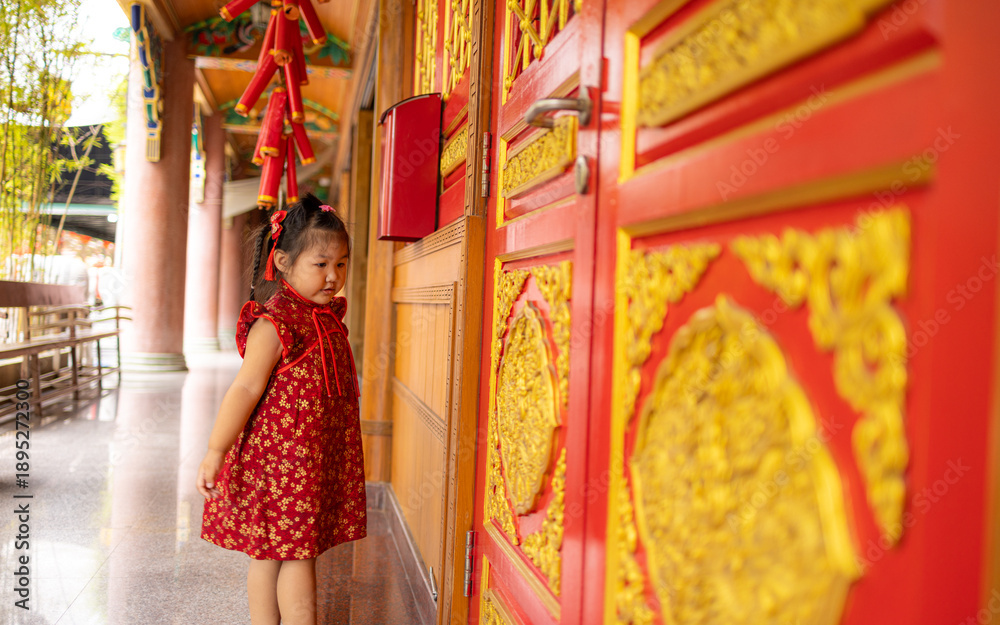Obraz premium Adorable Little Girl in Traditional Red Cheongsam Standing by Golden Carved Temple Doors,A cute young Asian girl wearing a festive red Chinese dress (Qipao) with gold floral patterns,