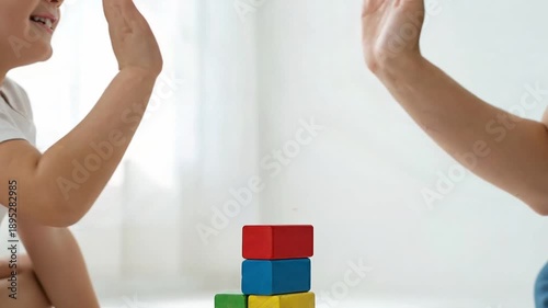 A close-up of a young boy and an adult building a tower with colorful wooden blocks. After finishing, they share a joyful high-five, celebrating teamwork, early childhood development, and success.